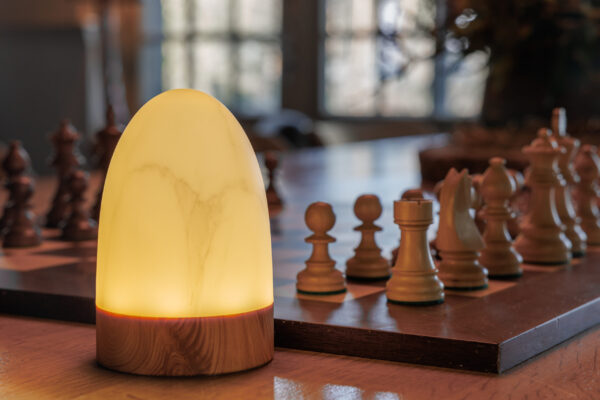 Een set van 2 ovale, waterdichte Tafellampen DORSETTO in marmer met grijs houtlook (oplaadbaar tot 105 uur, met afstandsbediening) staat op tafel naast een schaakbord in een zacht verlichte kamer met grote ramen.