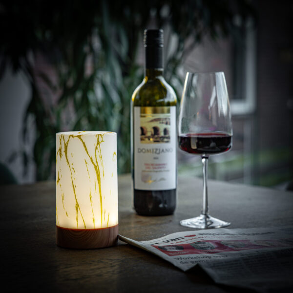 Een verlicht Tafellamp DORSETTO oplaadbaar 105 uur - waterdicht, cilindervorm, marmer goud houtlook, met afstandsbediening - staat op een tafel naast een wijnfles, glas en krant met groen en een onscherp raam op de achtergrond.