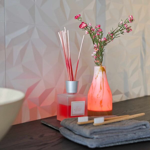 Een close-up van een aanrecht met twee tandenborstels, grijze handdoeken, een diffuser en de Tafellamp DORSETTO oplaadbaar 105 uur waterdicht ovaal marmer groen houtlook grijs goud met afstandsbediening die oplicht naast roze bloemen op een muur met structuur.