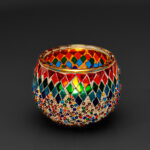 Theelichthouder gekleurde spikkels 9 CM DORSETTO: een ronde glazen kandelaar versierd met kleurrijke ruitvormige mozaïeken en veelkleurige kralen tegen een effen donkere achtergrond.