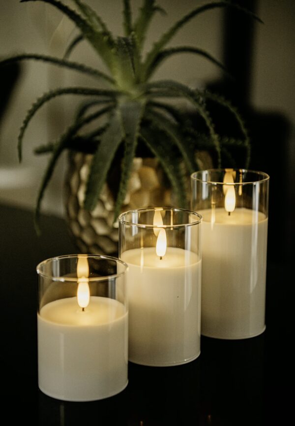 Drie DORSETTO led kaarsen met 3D vlam en oplaadbaar tot 105 uur staan in heldere glazen houders op een donker oppervlak, met een groene plant in een gouden pot onscherp op de achtergrond.