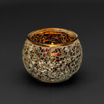 De theelichthouder 9 cm dorsetto beige bruin heeft een rond mozaïekglasontwerp in beige en bruin, dat warm opgloeit van een brandende kaars tegen een donkere achtergrond.