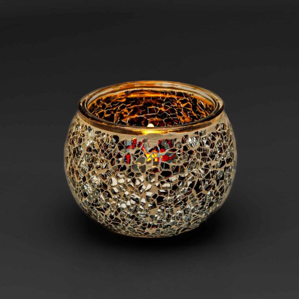 De theelichthouder 9 cm dorsetto beige bruin heeft een rond glazen ontwerp met een gouden en zilveren mozaïekpatroon, dat het licht prachtig weerkaatst om een warme, gloeiende sfeer te creëren tegen een donkere achtergrond.