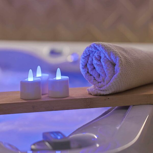 Een opgerolde beige handdoek en drie RGB LED-Waxine/theelichtjes (waterdicht, 68 uur oplaadbaar, set van 6 met afstandsbediening) worden op een houten plank boven een gloeiend bad geplaatst, waardoor een ontspannende spa-achtige sfeer wordt gecreëerd.