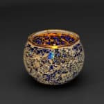 De Theelichthouder 9 cm DORSETTO donker blauw is een ronde glazen kandelaar met een blauw en gouden mozaïekontwerp, afgebeeld tegen een eenvoudige zwarte achtergrond.