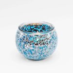 De Theelichthouder 9 CM Dorsetto lichtblauw is een ronde glazen theelichthouder met een mozaïek van lichtblauwe en zilveren scherven, waardoor een reflecterend en kleurrijk oppervlak ontstaat tegen een witte achtergrond.