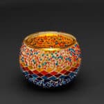 Theelichthouder 3 colormix 9 CM Dorsetto is een ronde kandelaar van mozaïekglas, versierd met rode, blauwe, gele en oranje kralen en tegeltjes. De levendige patronen weerkaatsen het licht prachtig tegen een donkere achtergrond.