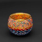 Theelichthouder 3 colormix 9 CM Dorsetto: een ronde glazen kandelaar versierd met levendige rode, blauwe, gele en oranje mozaïeksteentjes en kralen in decoratieve patronen, weergegeven tegen een donkere achtergrond.