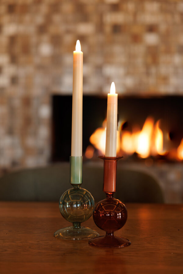 Twee kaarsen - een groene, een roodbruine - staan in glazen houders op een houten tafel. Daarachter gloeien een open haard en mozaĆÆektegels warm op, aangevuld met een Oplaadbare LED dinerkaars 270mm (130 uur) met afstandsbediening voor een moderne touch.