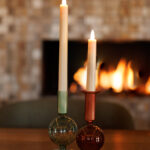 Twee Oplaadbare LED dinerkaarsen 270mm (warm wit, 130 uur, set van 12 met afstandsbediening) gloeien in gekleurde glazen houders op een houten tafel, met een zacht verlichte open haard erachter voor een gezellige sfeer.