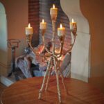 Een gouden, takvormige kandelaar met zes 3D oplaadbare LED-Waxine/theelichtjes (timerfunctie) staat op een houten tafel bij een brandende open haard en creëert een warme en gezellige sfeer.