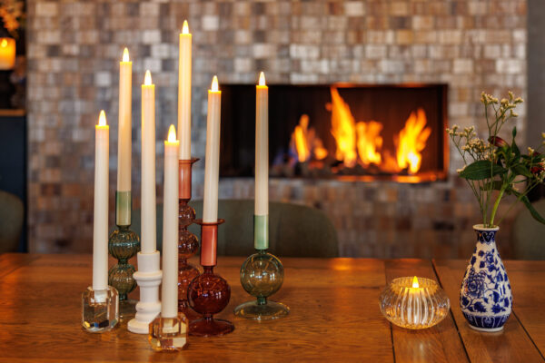 Een gezellig tafereel met brandende led dinerkaarsen in kleurrijke houders, bloemen in een vaas, een LED dinerkaars 270mm oplaadbaar 130 uur warm met 6 stuks met afstandsbediening op een houten tafel, en een gloeiend vuur in de open haard.