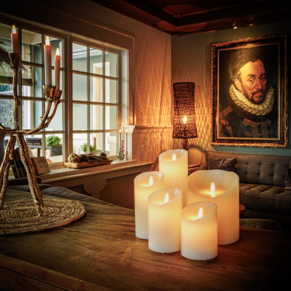 In een gezellige woonkamer gloeien de Oplaadbare LED waxinelichtjes D12,5 X H25 CM op een houten tafel, naast een kandelaar met kaarsen, een bank, een rieten lamp en een groot portret van een man. Warme sfeerverlichting vult de ruimte.