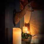 De LED kleine wax kaarsen set 2 kaarsen DORSETTO en feestelijk groen sieren een schouw onder een gemonteerde hertenkop en vullen de warm verlichte kamer met een gezellige, rustieke sfeer.