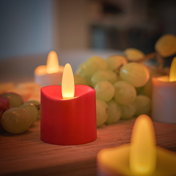 Een close-up van een gloeiend Rode oplaadbaar LED waxine kaarsje (175 uur, waterdicht, met afstandsbediening, timerfunctie), naast groene druiven op hout, met andere zacht brandende kaarsen uit de 6-stuks set op de achtergrond.