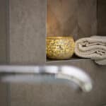 Een Theelichthouder zwart 9 CM DORSETTO staat op een badkamerplankje naast netjes gevouwen crèmekleurige handdoeken, met een vage kraan zichtbaar op de voorgrond.