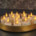 Een rond gouden dienblad toont 12 3D oplaadbare LED-Waxine/theelichtjes (175 uur, met afstandsbediening, timerfunctie), hun kunstmatige vlammen dicht bij elkaar geplaatst voor een warme, gezellige sfeer tegen donkere tegels.