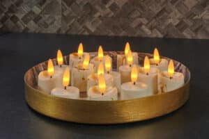 Een rond gouden dienblad bevat verschillende led waxinelichtjes 3D brandtijd 175 uur oplaadbaar - met Afstandsbediening - 6 Stuks - timerfunctie, gerangschikt op een donker aanrecht met een tegelachterwand met textuur op de achtergrond.