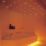 Een modern bad in een zacht verlichte badkamer straalt met warm oranje licht. Het plafond is voorzien van een LED Sterrenhemel RGB hoge kwaliteit (180 sterren) met afstandsbediening, die sterreneffecten werpt voor een ontspannen, luxueuze sfeer. Handdoeken en decor liggen op de rand van het bad.