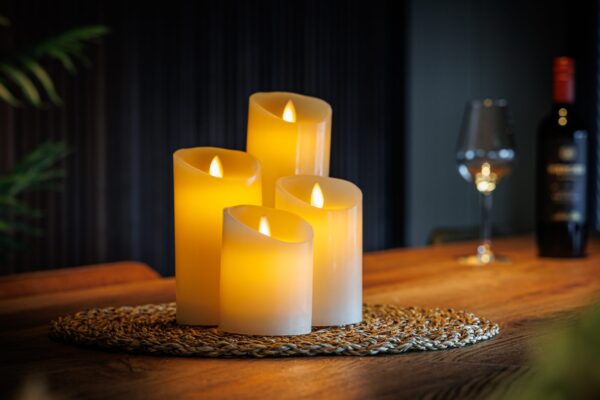 Een set van vier Oplaadbare LED wax kaarsen van 4 DORSETTO met verschillende hoogtes ligt op een geweven mat boven op een houten tafel en verlicht zacht de achtergrond waar een wijnglas en een fles de warme sfeer versterken.