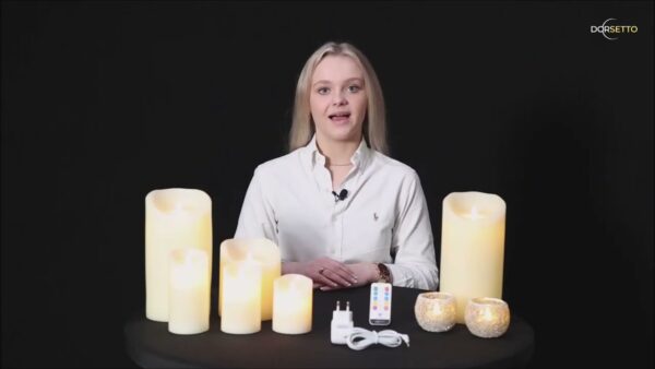 Een vrouw in een wit shirt zit aan een tafel met de DORSETTO LED wax kaarsen set 4 kaarsen warm met oplaadbaar, afstandsbediening en voedingsadapter tegen een zwarte achtergrond.