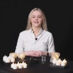 Een vrouw met lang blond haar zit aan een ronde zwarte tafel gedekt met brandende kaarsen, waaronder Dorsetto Oplaadbare LED waxine/theelichtjes (6 stuks, 175 uur, waterproof, warm wit, met afstandsbediening), tegen een zwarte achtergrond. Ze glimlacht naar de camera.