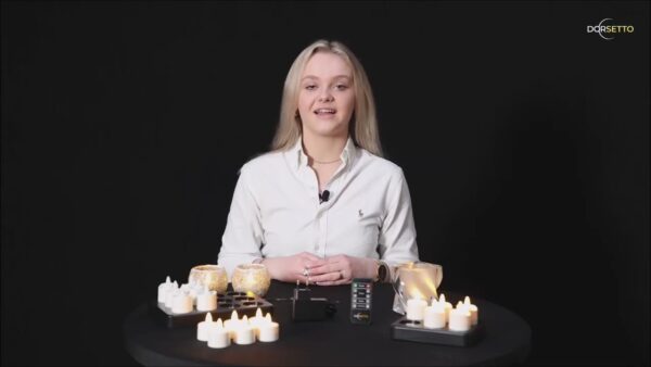 Een vrouw met lang blond haar zit aan een ronde zwarte tafel gedekt met brandende kaarsen, waaronder Dorsetto Oplaadbare LED waxine/theelichtjes (6 stuks, 175 uur, waterproof, warm wit, met afstandsbediening), tegen een zwarte achtergrond. Ze glimlacht naar de camera.