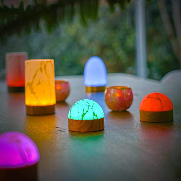Kleurrijke decoratieve lampen, waaronder de waterdichte Tafellamp DORSETTO oplaadbaar 105 uur in marmergroene houtlook met afstandsbediening, gloeien op een houten oppervlak tegen een vage achtergrond buiten.