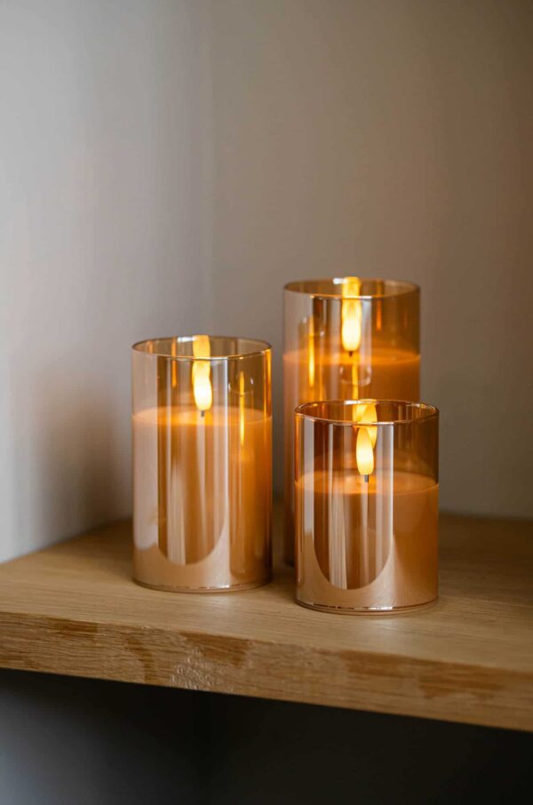 Drie DORSETTO oplaadbare led kaarsen in amberkleurige houders, 105 uur brandtijd en afstandsbediening, staan op een houten plank en geven sfeervol licht. Het magnetisch oplaadstation kleurt rood tijdens het opladen van de 3-delige set.