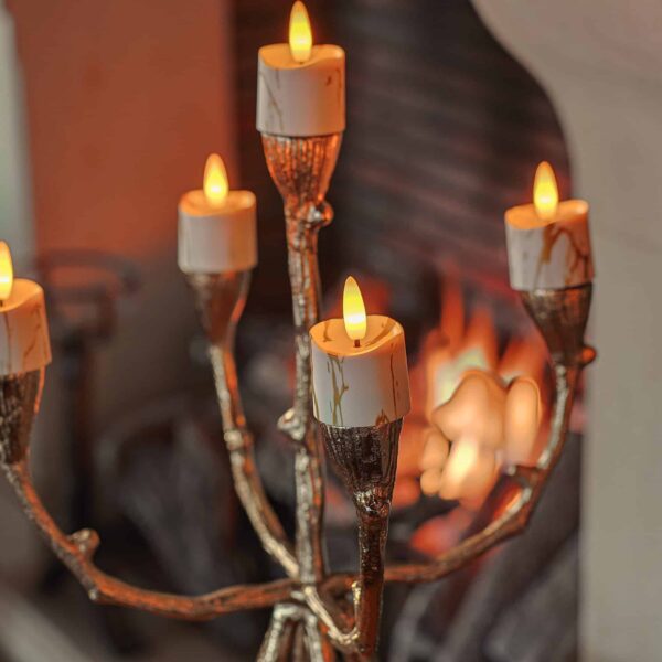 Een gouden kandelaar met vijf 3D oplaadbare LED-waxine/theelichtjes (set van 12, timerfunctie, afstandsbediening) staat voor een open haard. Het takvormige ontwerp en de gloeiende lampjes creëren een gezellige, warme sfeer.