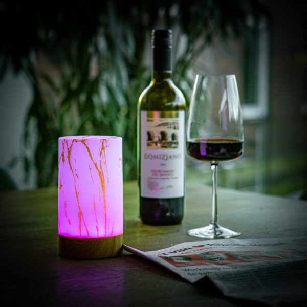 De oplaadbare Tafellamp DORSETTO (105 uur, waterdicht, cilindervorm, marmer wit-grijs houtlook, met afstandsbediening) staat op een tafel naast een wijnfles, glas en krant, met groen en ramen op de achtergrond.