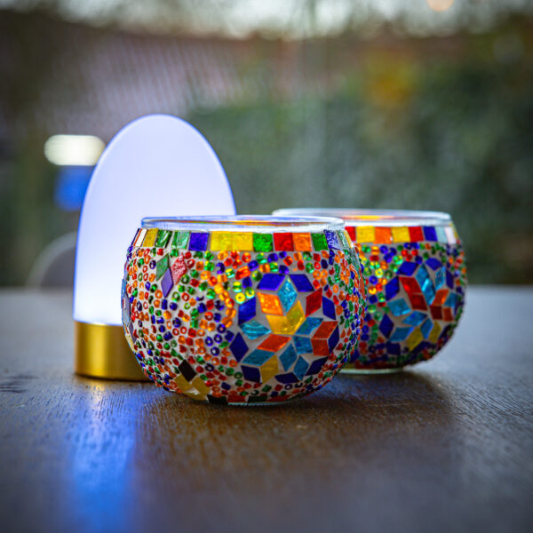 Twee kleurrijke kandelaars van mozaïekglas zitten naast de Tafellamp DORSETTO oplaadbaar 105 uur - een ovale, wit-gouden, waterdichte LED-lamp met afstandsbediening - die zachtjes op een houten oppervlak brandt. De achtergrond is onscherp.