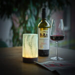 Op een houten tafel staan de oplaadbare Tafellampen DORSETTO (set van 2, 105 uur, waterdicht, cilindervorm groen marmer houtlook met afstandbediening), samen met een fles rode wijn, een gevuld wijnglas en een opgevouwen krant. Een groene plant is vaag op de achtergrond.
