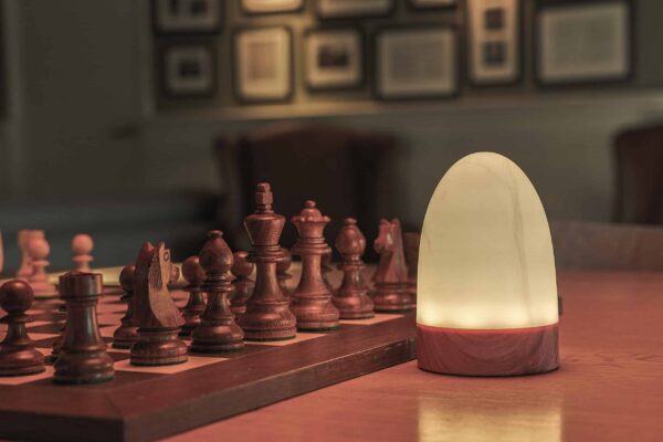In een warm verlichte kamer staat de tafellamp DORSETTO oplaadbaar 105 uur, waterdicht en ovaal met marmer met grijze houtlook en afstandsbediening, te stralen naast een houten schaakbord met uitgesneden stukken op tafel.