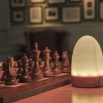 A softly glowing Tafellamp DORSETTO oplaadbaar 105 uur, waterdicht, ovaal met marmer groen houtlook grijs goud en afstandsbediening staat op een houten tafel naast een schaakbord; vage foto's en documenten hangen op de achtergrond aan de muur.