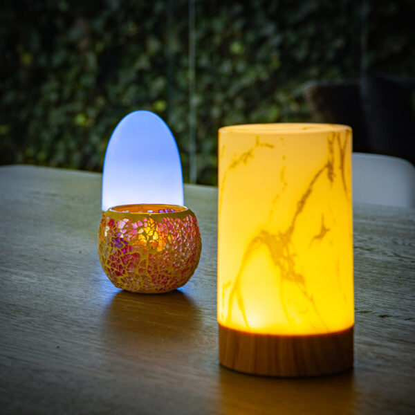 Drie decoratieve lampen rusten op een houten tafel: een lamp met geel marmerdessin aan de voorkant, een lamp met goudstructuur en paars licht zoals de Waxinelichthouder 8,5 CM Dorsetto mozaïekroos in het midden, en een blauwgloeiende lamp aan de achterkant, met groen erachter.