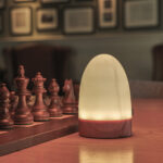 Een set van twee waterdichte led Tafellampen DORSETTO oplaadbaar 105 uur, ovaal met afstandsbediening en marmer wit-grijs houtlook, staat naast een houten schaakset op de tafel, met vage fotolijsten op de achtergrond.