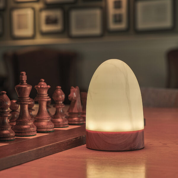 Een set van twee waterdichte led Tafellampen DORSETTO oplaadbaar 105 uur, ovaal met afstandsbediening en marmer wit-grijs houtlook, staat naast een houten schaakset op de tafel, met vage fotolijsten op de achtergrond.