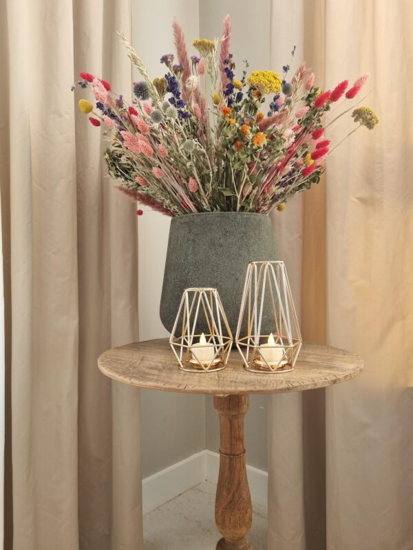 Een rustieke houten tafel met een grote vaas met kleurrijke droogbloemen en een Waxinelichthouderset DORSETTO metaal rosé goud kleurig per 2 stuks, elk met brandende waxinelichtjes, is gedekt tegen beige gordijnen.