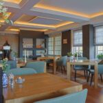 Modern restaurantinterieur met houten tafels, groene stoelen met kussens, grote ramen met vouwgordijnen, zachte plafondverlichting, potplanten, decoratieve vazen en LED waxinelichtjes 3D (175 uur brandtijd, oplaadbaar) voor een gezellige sfeer.