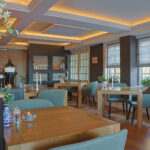 Een stijlvol restaurant met houten tafels, groene stoelen, zachte verlichting van LED dinerkaars 270mm oplaadbaar 130 uur warm wit (6-pak met afstandsbediening), grote ramen met vouwgordijnen, bloemen, kaarsen, vazen en uitgestalde wijnflessen.