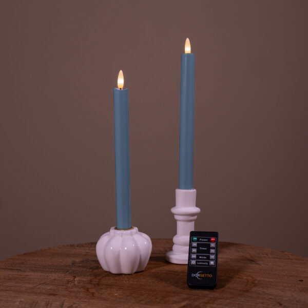 Twee Dinerkaarsen LED kaarsen (steenblauw, set van 2, 400 uur oplaadbaar) in witte houders staan op een houten ondergrond met een zwarte afstandsbediening ervoor; effen bruine achtergrond.