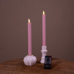 Two Dinerkaarsen LED 400 uur brandduur oplaadbaar in zacht roze, set van 2, staan in witte houders op een houten oppervlak. Een kleine afstandsbediening ligt ervoor. De achtergrond is effen bruin.