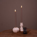 Twee beige Dinerkaarsen LED kaarsen (set van 2, 400 uur oplaadbaar) staan in witte houders op een houten oppervlak, met een kleine zwarte afstandsbediening ervoor. De effen bruine achtergrond maakt het tafereel compleet.