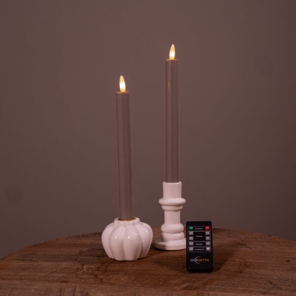Twee beige Dinerkaarsen LED kaarsen (set van 2, 400 uur oplaadbaar) staan in witte houders op een houten oppervlak, met een kleine zwarte afstandsbediening ervoor. De effen bruine achtergrond maakt het tafereel compleet.