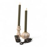 Twee groene kaarsen met crèmekleurige houders, een zwarte afstandsbediening en een zwarte stroomadapter staan bij elkaar op een witte achtergrond. De kaarsen lijken elektrisch te zijn met vlamvormige uiteinden.