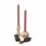 Twee roze kaarsen op batterijen in witte houders, een afstandsbediening en een zwarte voedingsadapter staan op een witte achtergrond. De kaarsen hebben vlamvormige uiteinden die echte vlammen simuleren.