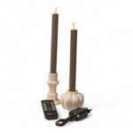 Twee bruine vlamloze kaarsen in witte houders, vergezeld van een zwarte afstandsbediening en een stroomadapter, worden weergegeven tegen een effen witte achtergrond.